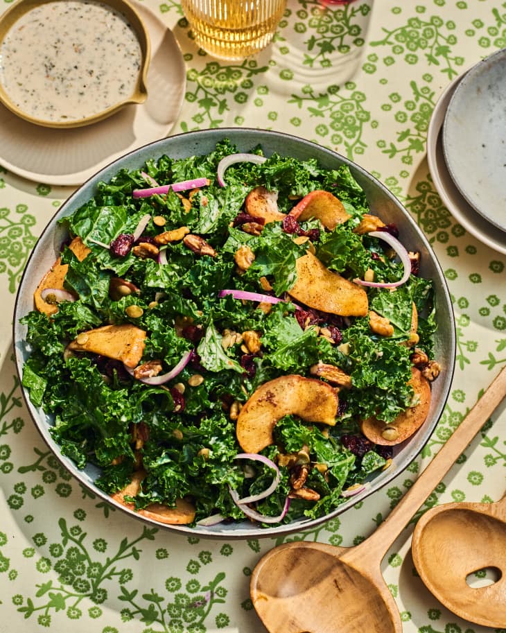 kale and roasted apple fall salad on thanksgiving table. there are wooden salad spoons in the shot, and a small bowl of dressing in the upper left. floral green and off-white tablecloth