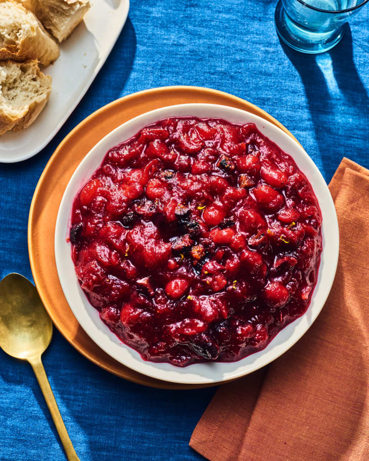 bowl of cranberry sauce on yellow plate on thanksgiving table. there is a blue tablecloth, a terracotta colored napkin, and a gold spoon. There as a plate of rolls peeking into frame in the top left corner