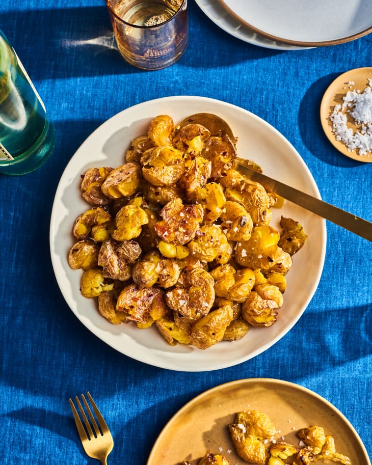 Serving bowl of smashed potatoes with serving spoon on table. Bright blue tablecloth, some kosher salt in a small bowl, a plate with a serving of potatoes, and a bottle of wine and a glass of wine peeking in