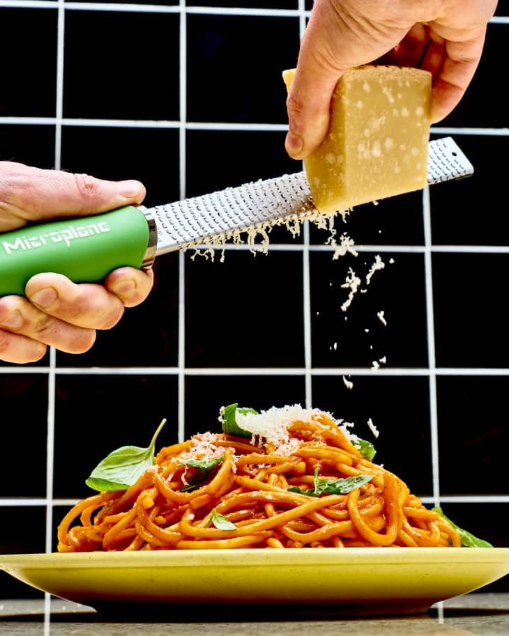 Hands grating parmesan cheese with a microplane zester over a heaping plate of spaghetti with red sauce. Black tile background