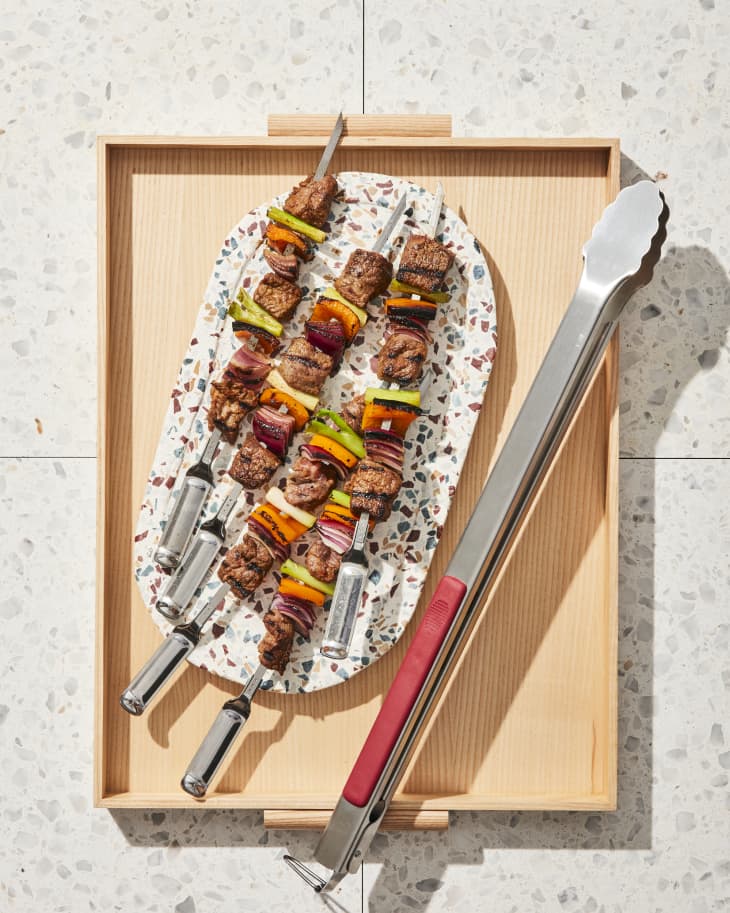 wood tray on countertop  with speckled plate of grilled beef, pepper, and onion skewers. Pair of tongs on tray as well