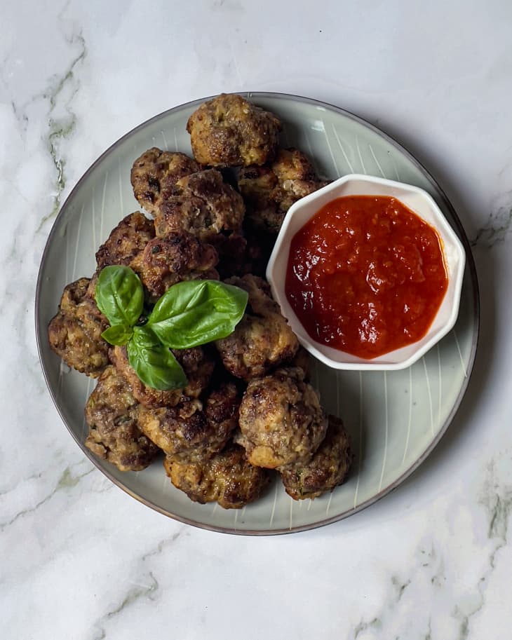 15 small meatballs sitting on a gray round plate with red sauce in a small round bowl on the plate, with three basil leafs on top