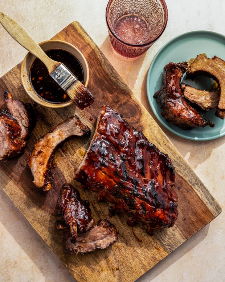 A rack of baby back ribs on a wooden platter, with bbq sauce and a brush also on the platter and a plate with ribs nearby
