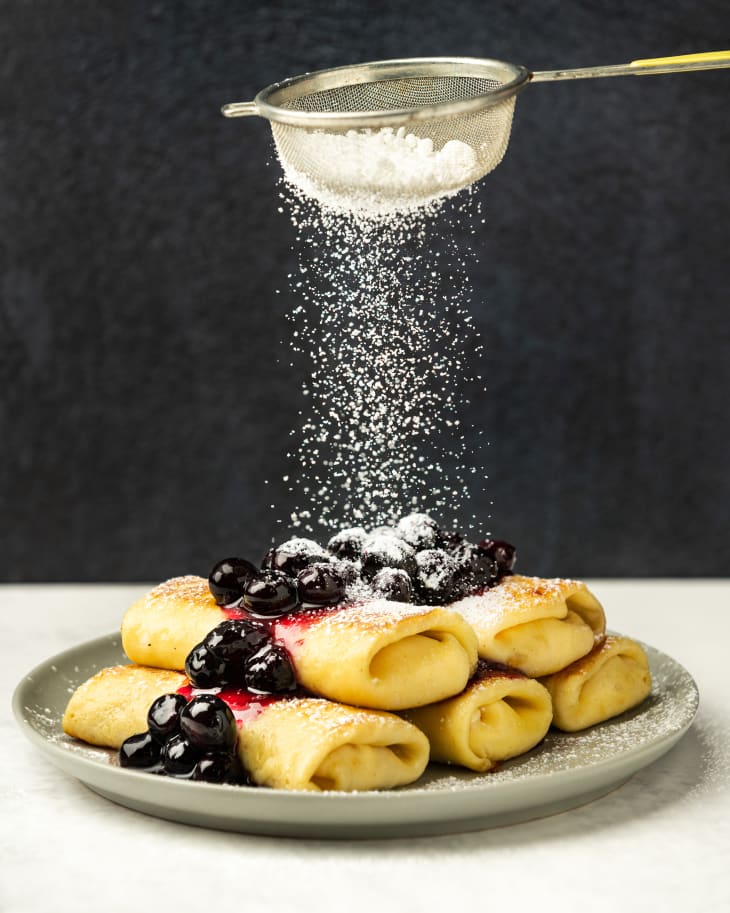 A stack of five cheese Blintz's on a plate, with blueberry sauce on top, and powdered sugar being shaken over the top.