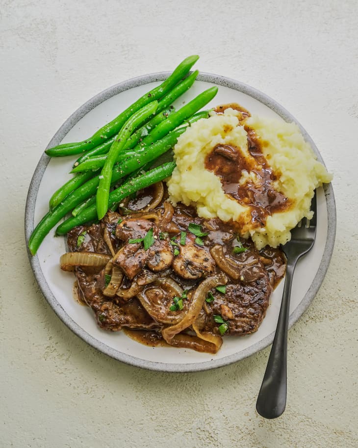 Cube Steak, which is ground beef cooked with gravy and vegetables, on a plate mashed potatoes and gravy, and green beans as side dishes. There is a fork on the plate