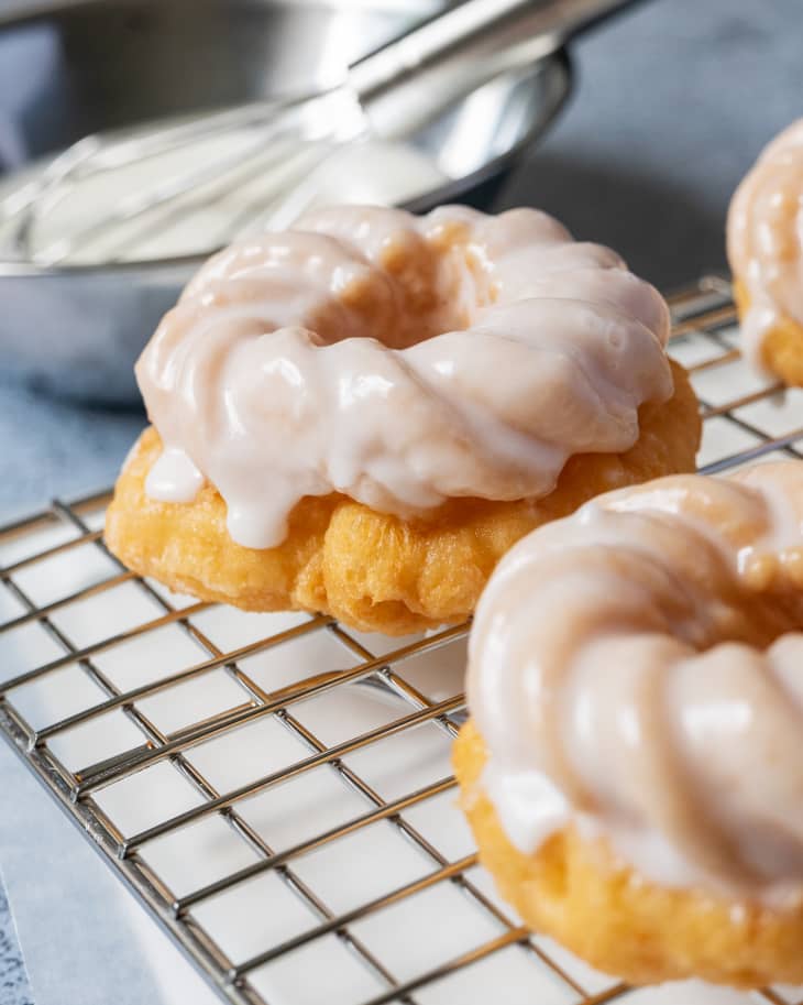 a donut with white glaze on a cooling rack, with a whisk in a bowl of glaze in the background