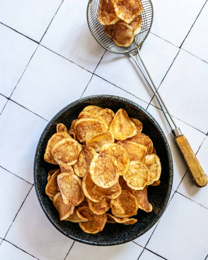 A bowl of lightly browned homemade potato chips on a white tiled countertop, with a strainer with some more chips next to it