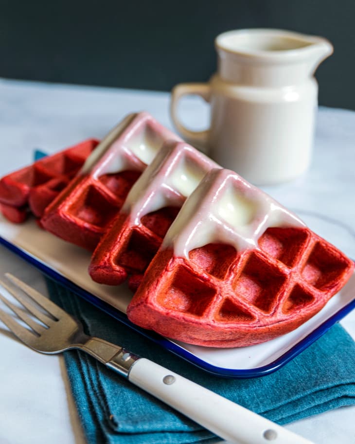 Red Velvet Waffle cut into four pieces, with the corners dipped into white icing with a fork, blue napkin and white pitcher with syrup next to it.