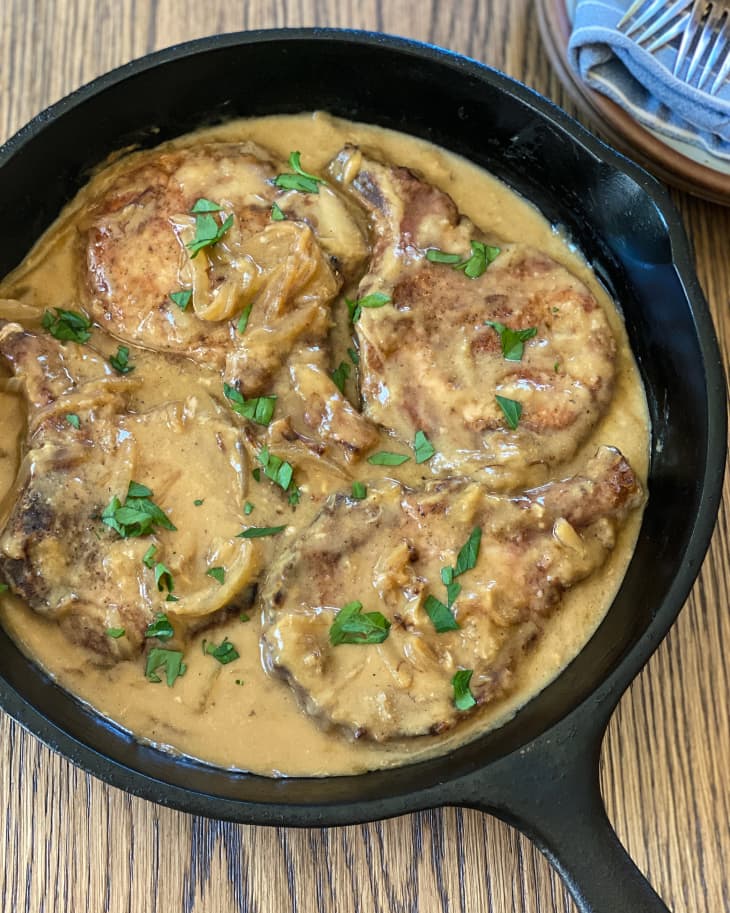Four Smothered Pork Chops in a black skillet with brown gravy and and green garnish on top.