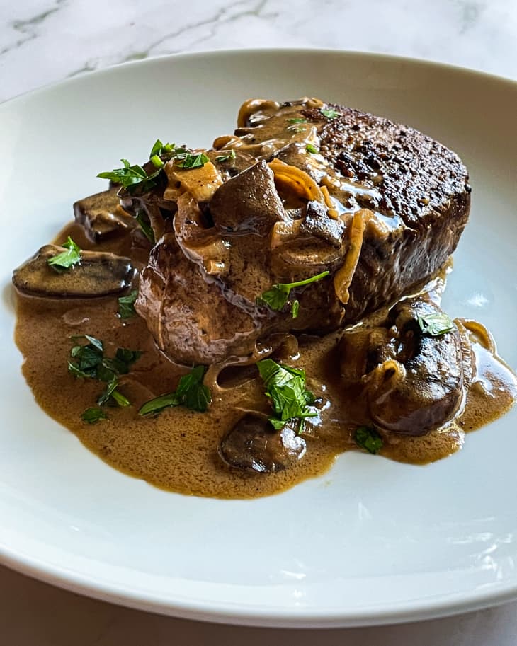 Steak Diane on a white plate; a piece of beef steak, seared, with brown gravy and crimini mushrooms on top. There is also a green parsley garnish.