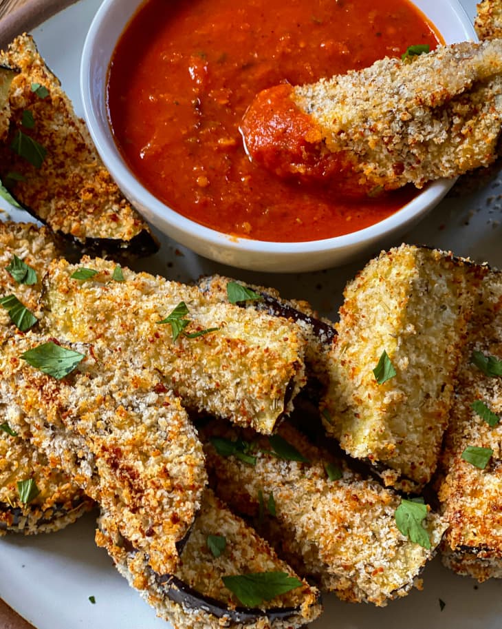 Air Fryer Eggplant on a plate with a small bowl of marinara sauce on the side and a green garnish sprinkled on top, with one fried eggplant stick being dipped into the marinara.