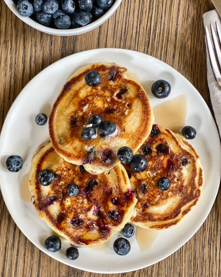three blueberry pancakes on a plate with extra blueberries sprinkled on top and in a bowl beside the plate.