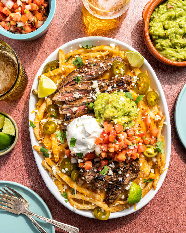 french Fries with Carne Asada beef. guacamole, salsa, sour cream, lime wedges and crumbled white cheese on top, surrounded by plates with the aforementioned ingredients.