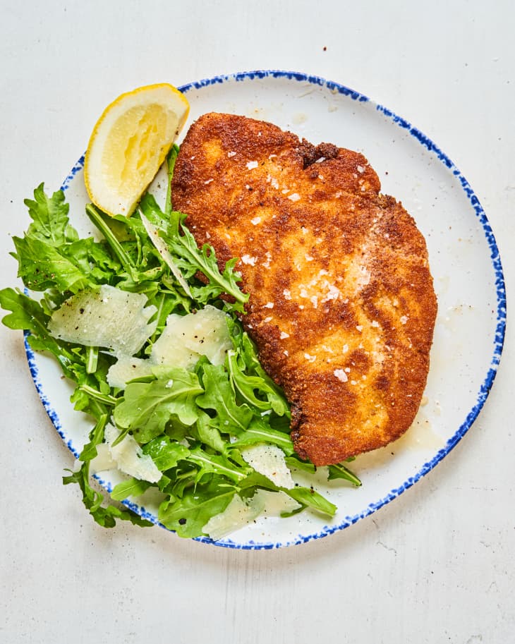 Chicken Milanese, on a plate with greens with shaved parmesan cheese and a lemon wedge.