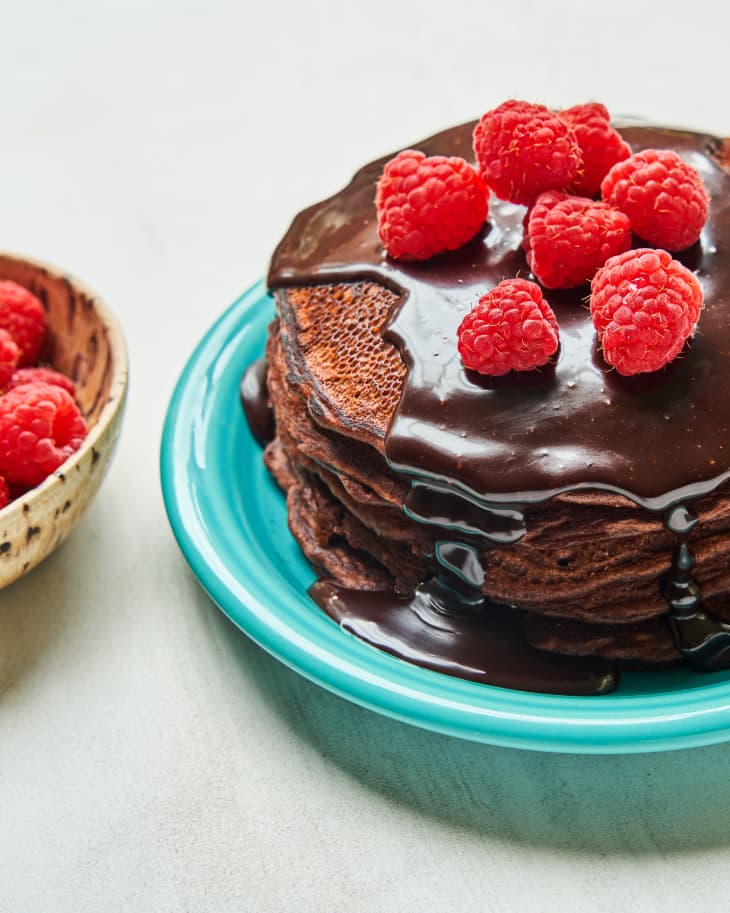 a stack of chocolate pancakes on a plate with chocolate sauce and raspberries on top, and a bowl of raspberries off to the side.