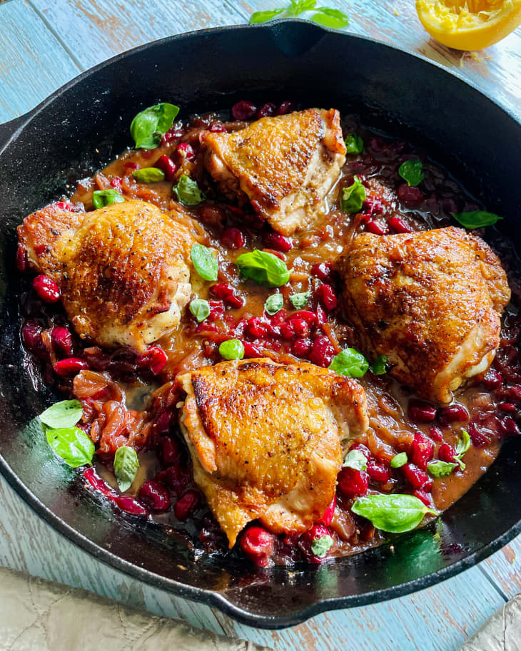 Cranberry chicken in a cast iron pan, on a wooden tabletop with a burlap napkin next to it.