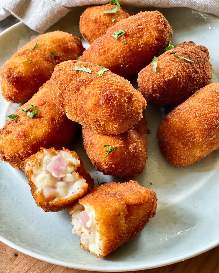 A pile of croquetas on a white plate with a green garnish, with one Croqueta broken in half to show the ham and cheese inside.