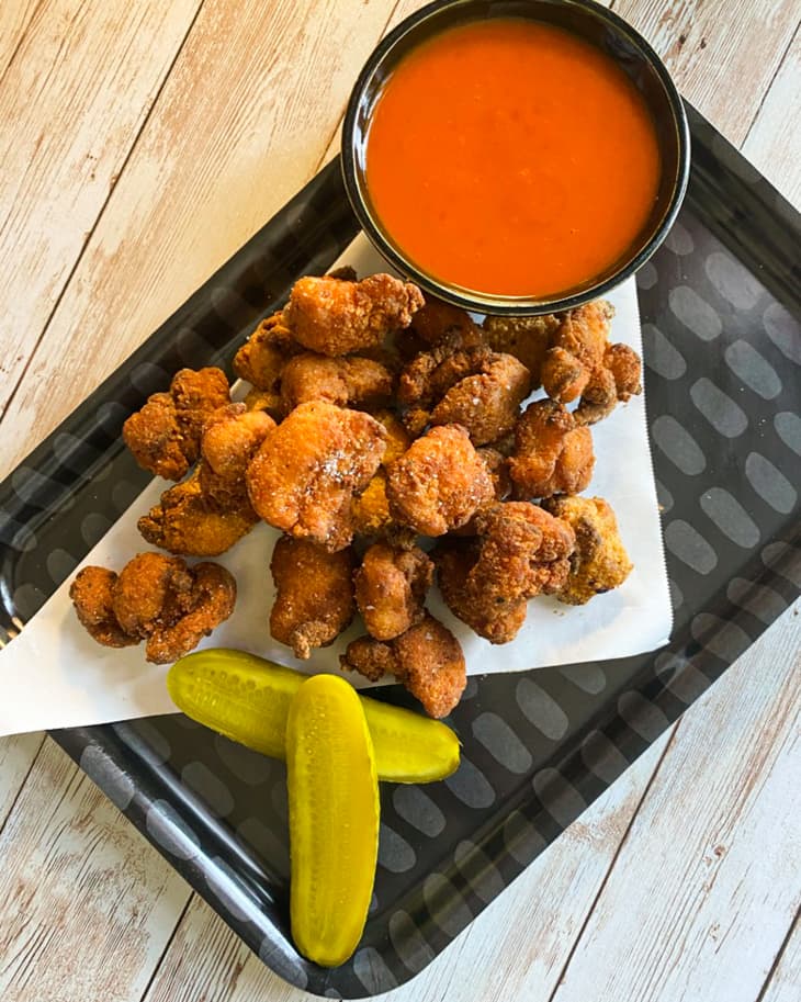 popcorn chicken (small pieces of fried chicken) on a tray with a pickle cut in half and a ramekin of red sauce.