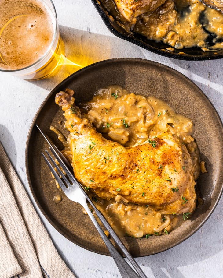 smothered chicken on a plate with utensils next to it, a glass of beer and the pan next to it