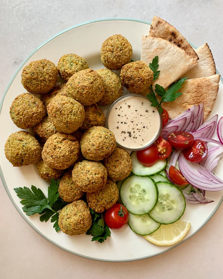 a plate full of air fryer falafel balls with dipping sauce, pita bread, onions, cucumbers, tomato and parsley