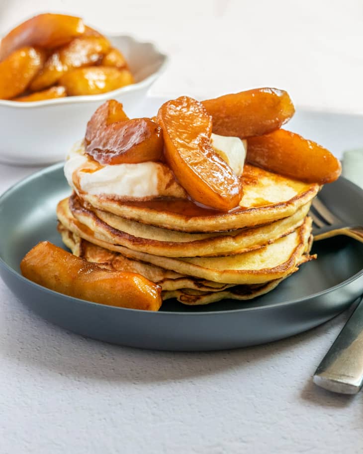 A stack of pancakes, topped with caramelized, baked apple slices, on a blue plate, with a white bowl more apples  slices in the background.