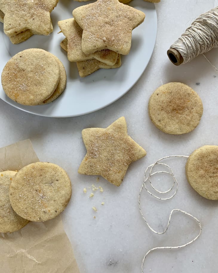 Biscochitos (Mexican biscuits or cookies) cut into circle and star shapes, spread across a white plate and also across a marble countertop, with bakery string laying nearby