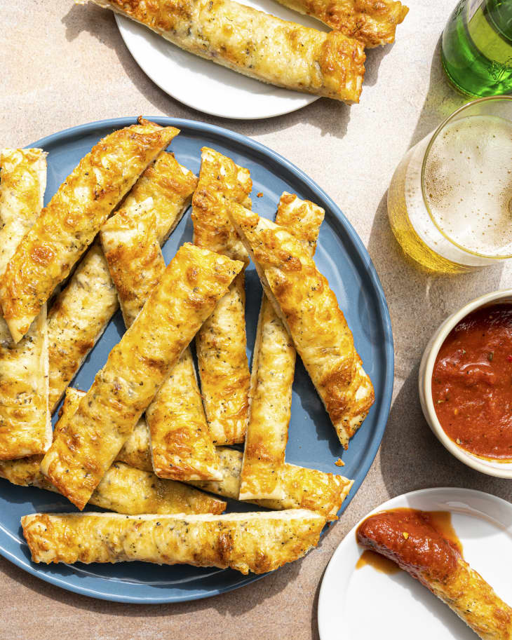 A pile of cheese breadsticks on a round blue plate, with a side of marinara sauce, a small plate to the side with a single cheesy breadstick dipped in sauce, a glass of beer and another plate with more breadsticks nearby.