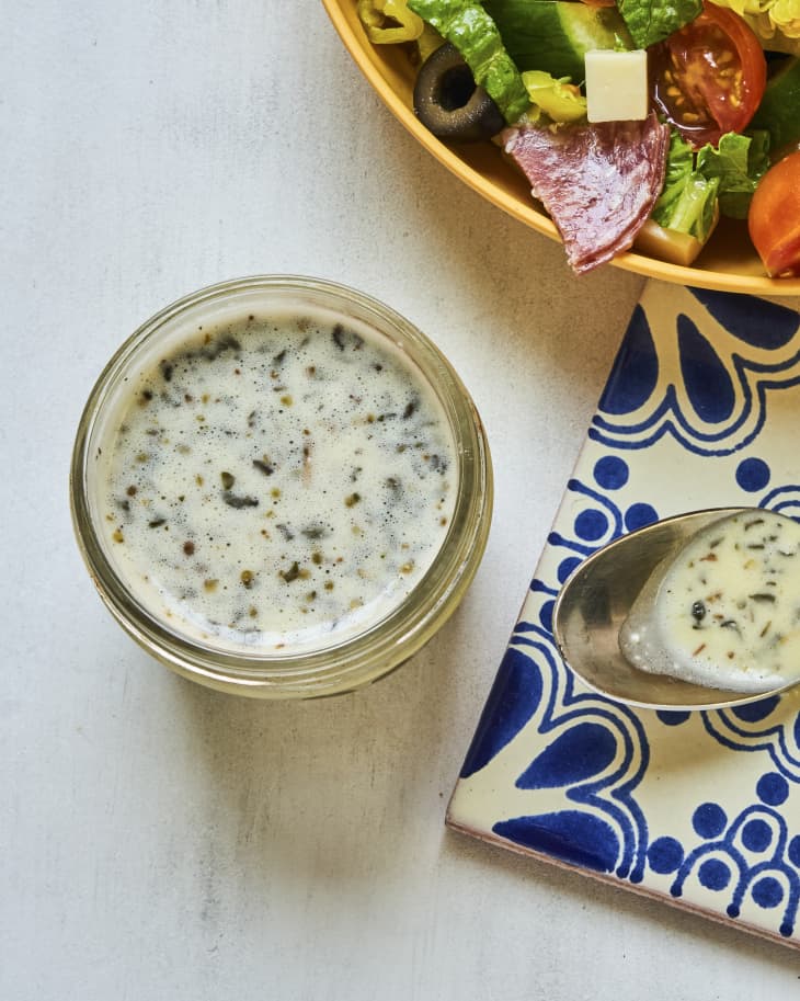 Creamy Italian dressing in a small open jar, with an Italian style salad with cucumbers, olives, tomatoes, cheese and cured meats on the side, and a plate with a blue and white design with a spoon with more dressing on it on the the side.