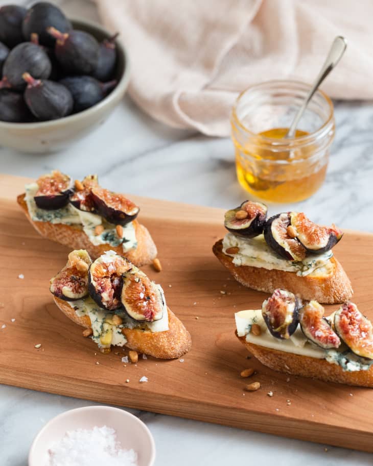 Crostini (crusty bread brushed with olive oil, sliced and toasted in the oven or in the grill), on a wooden cutting board with toppings, with olive oil and figs in a bowl in the background