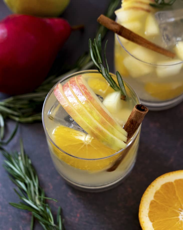 Sangria with apples, oranges, amd spices in a clear glass with a cinnamon stick, with another sangria glass nearby, and a part of a whole apple, and orange cut in half also nearby.
