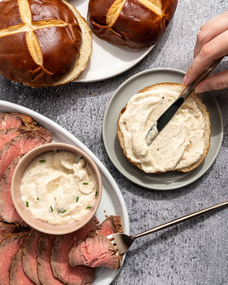 Horseradish sauce being spread onto a roll, with a plate of roast beef and a bowl of horseradish sauce next to it, and a two sliced pretzel rolls to the side.