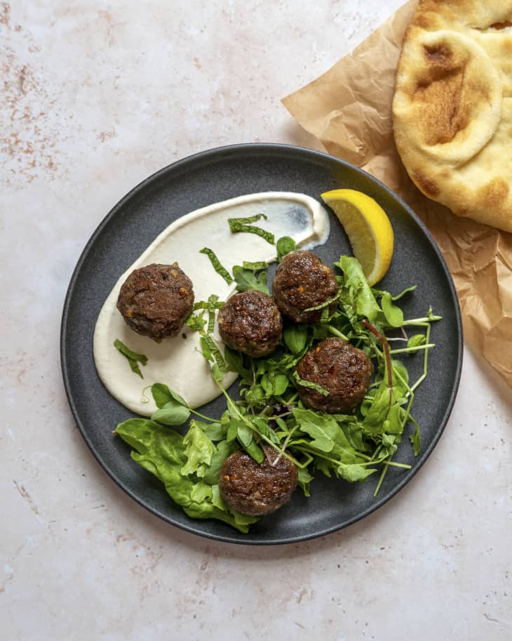Lamb meatballs on a black plate with white sauce and greens, and a lemon wedge.