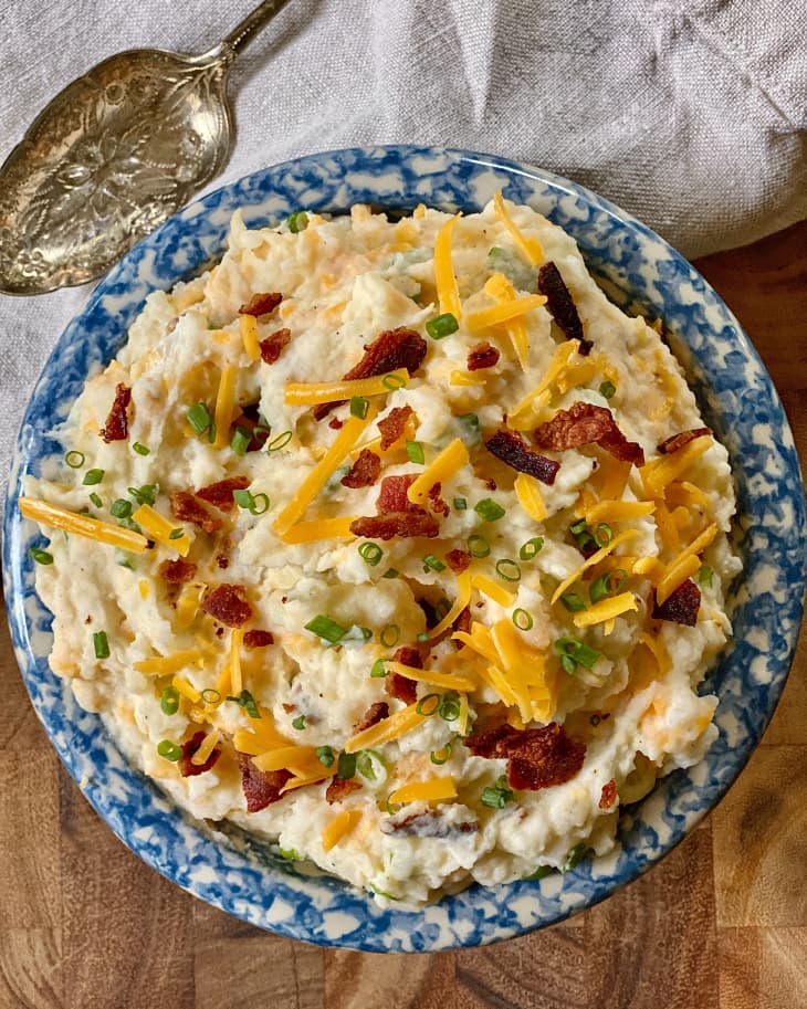 Loaded Mashed Potatoes (mashed potatoes topped with grated cheddar cheese, bacon, green onion) in a blue and white bowl, with a napkin and silver spoon beside it.