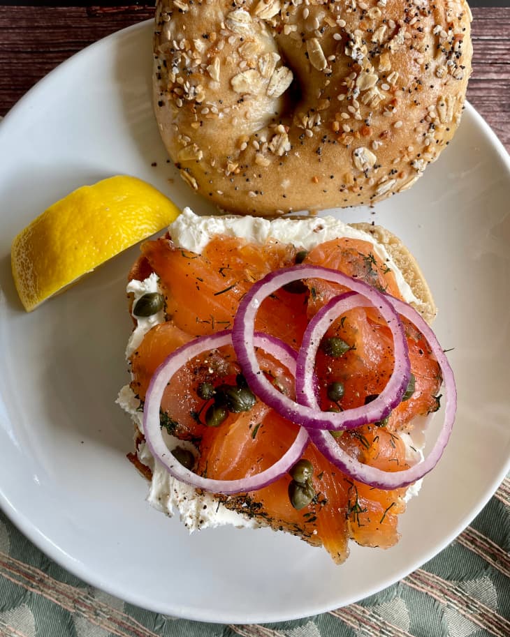 A top down look at an everything, multigrain bagel cut in half, with lox, sliced red onion, capers and creamcheese on one side, and the other half of the bagel with seeds on the other side of the plate. There is also a lemon wedge on the plate.
