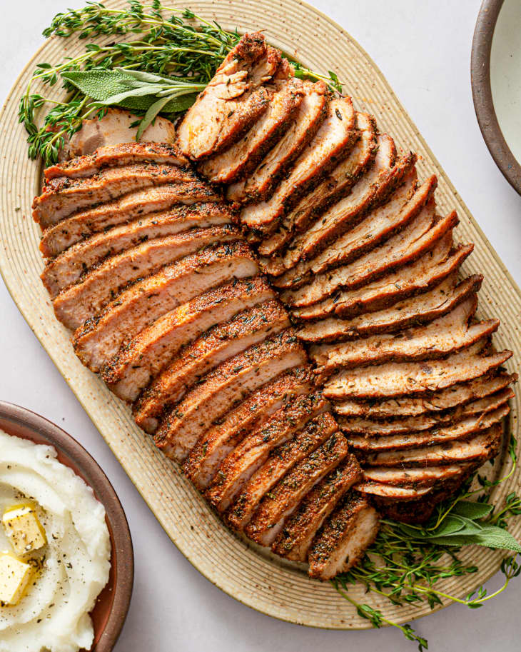 Turkey Tenderloin, roasted and sliced in two rows, on a rounded wooden platter with a rosemary and sage garnish