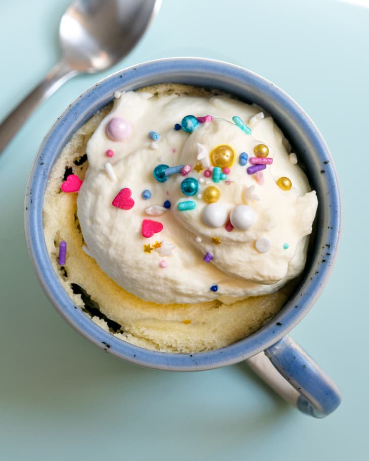 Vanilla cake baked in a blue mug, with whipped cream and colored sprinkles on top. There is a spoon on the side.