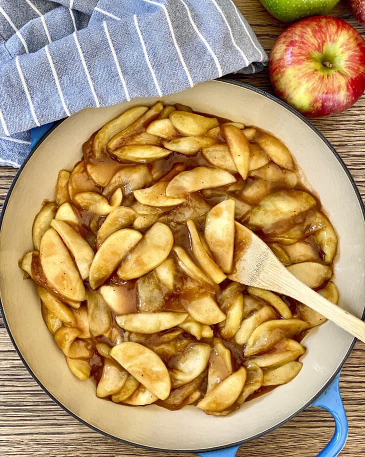 Apple Pie filling in a tan ceramic bowl with a thin black rim, with a wooden spoon in, and a blue and white striped napkin above, as well as an apple