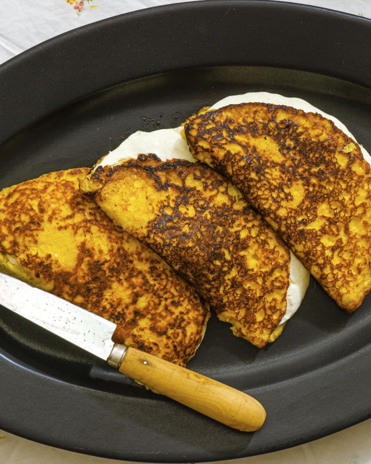 three Cachapa's (Venezuelan corn cakes) folded in half with creamy white cheese inside, on a black plate, with a knife on the side
