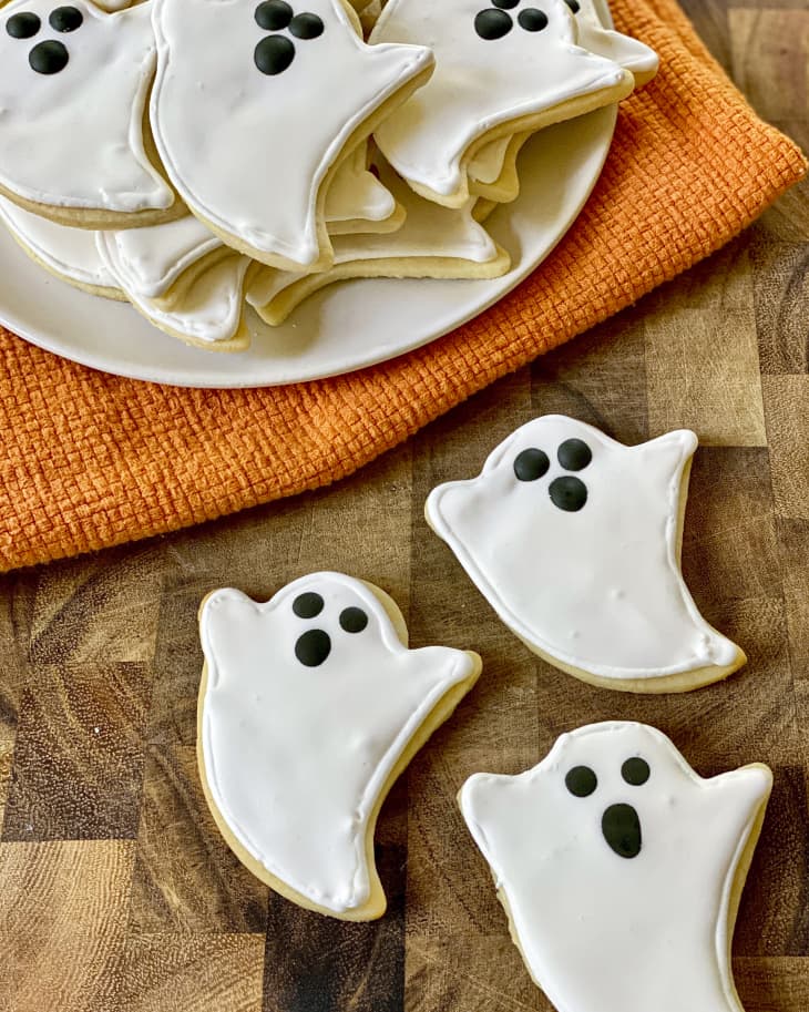 Three sugar cookies in the shape of ghosts, with white icing for the body and black icing for the face are on a wooden surface. There are a pile of cookies on a plate behind them, sitting on an orange cloth.