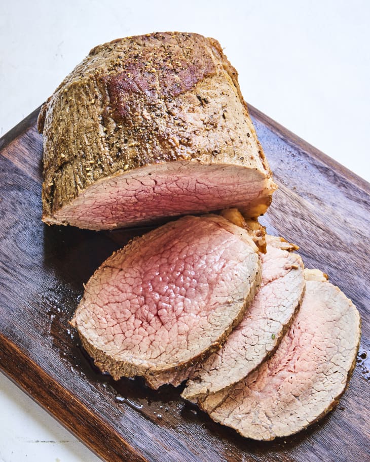 a cut of roast beef on a wooden cutting board with three slices laying on their sides.