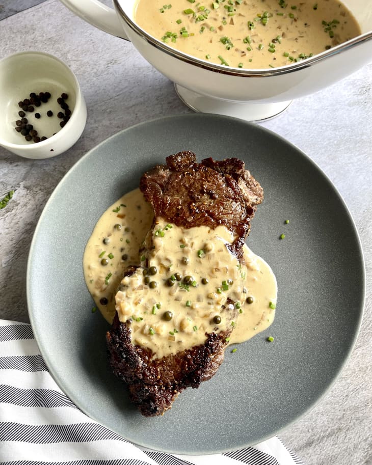 Creamy peppercorn sauce poured over a steak, on a gray-blue playe, with a gray and white striped napkin, a bowl of the peppercorn sauce in the background, and a small white ramekin with whole peppercorns in it.