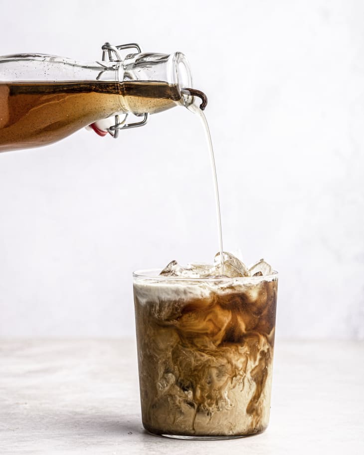 A glass cup with coffee and cream barely mixed together so it appears marbleized, as vanilla syrup is being poured