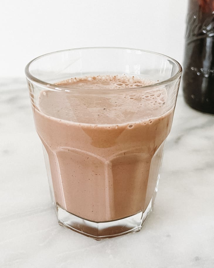 Chocolate milk in a glass, with a ball jar with chocolate sauce in the background, on a marble surface.