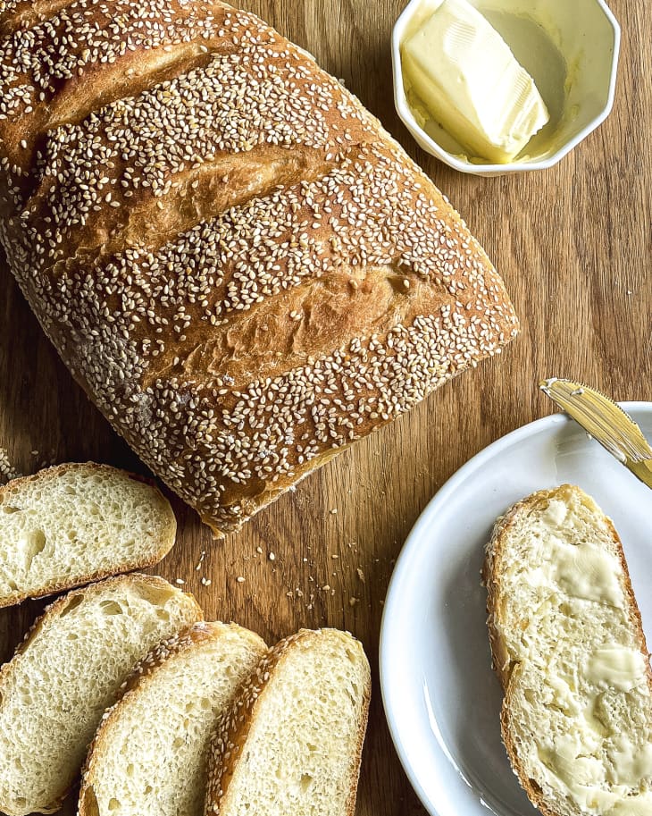 Homemade Italian bread with sesame seeds, with some slices cut off, and a slice with butter on a white plate on the side