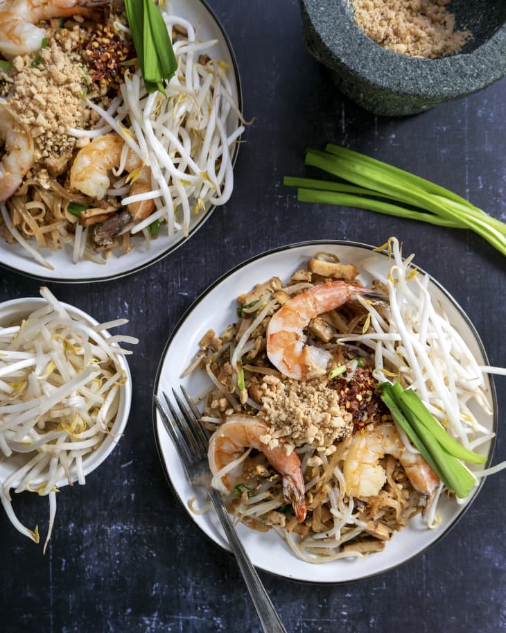 two plates of pad thai with shrimp, beansprouts, and green onions, with more of the garnishes in plates on the side.