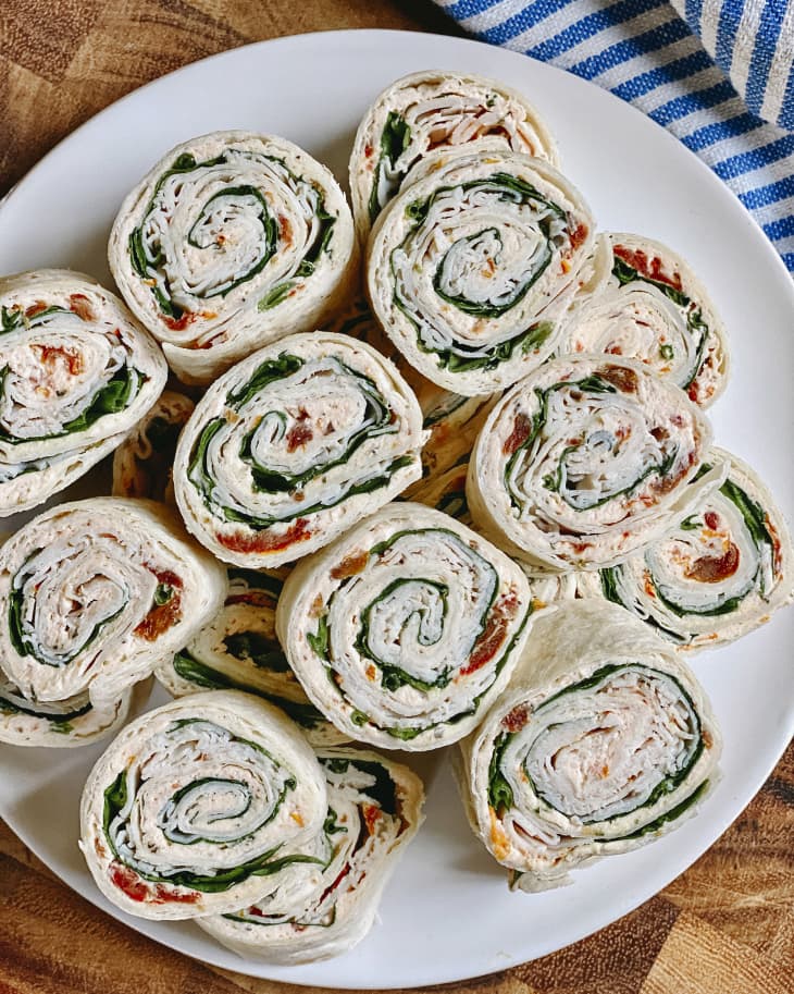 a stack of Pinwheels (tortillas wrapped with various ingredients and sliced like sushi) on a white plate with a blue striped napkin behind it.