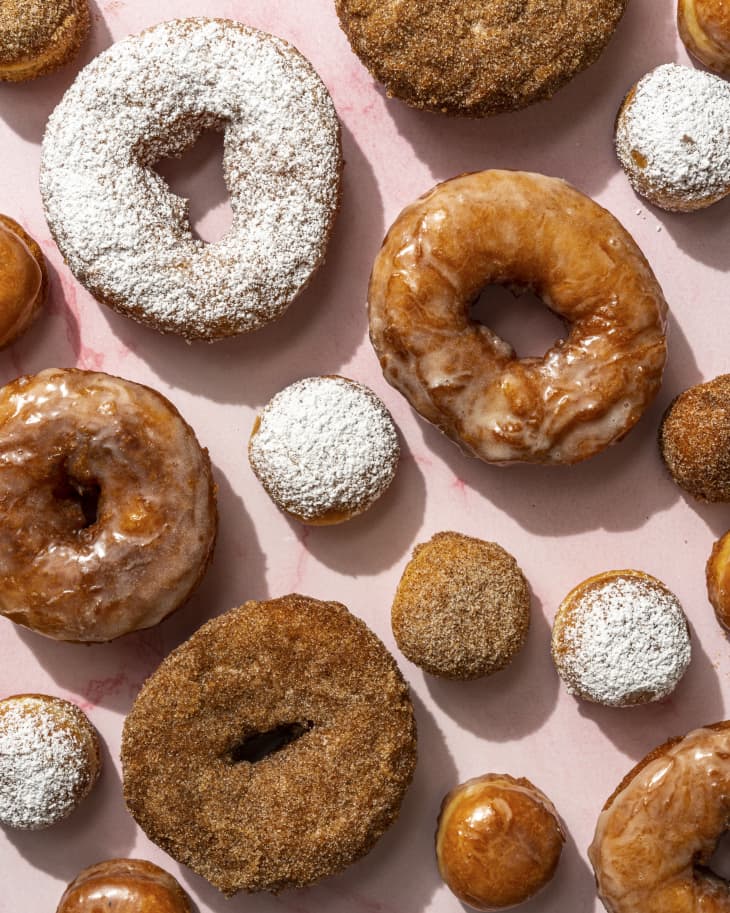 donuts of different shapes and sizes with various glazes and sugars arranged on a pink background.