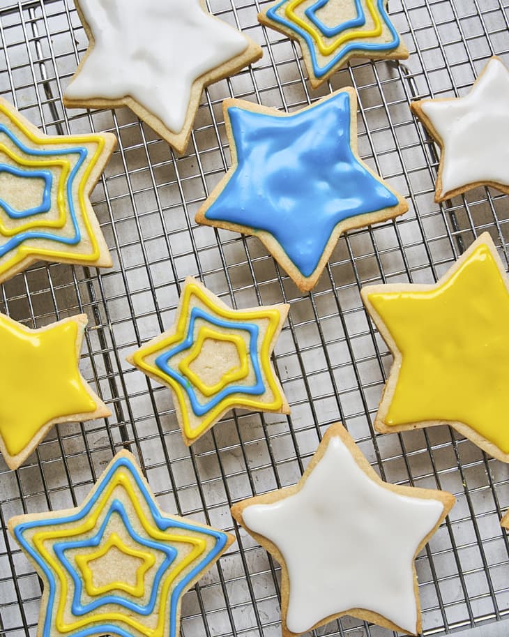 sugar cookies in the shape of stars with blue, yellow and white icing decorating the cookies in various designs, with some of the cookies iced in a single, solid color, on a cooling rack