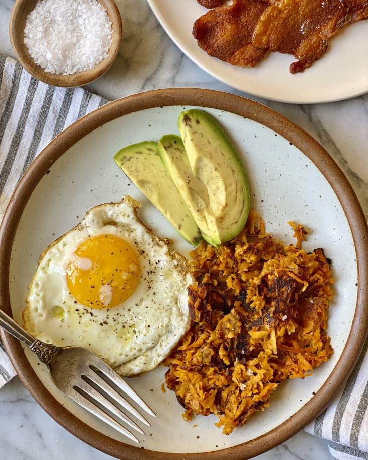 sweet potato hashbrowns on a plate with a fried egg and avocado, and bacon on a plate on the side.