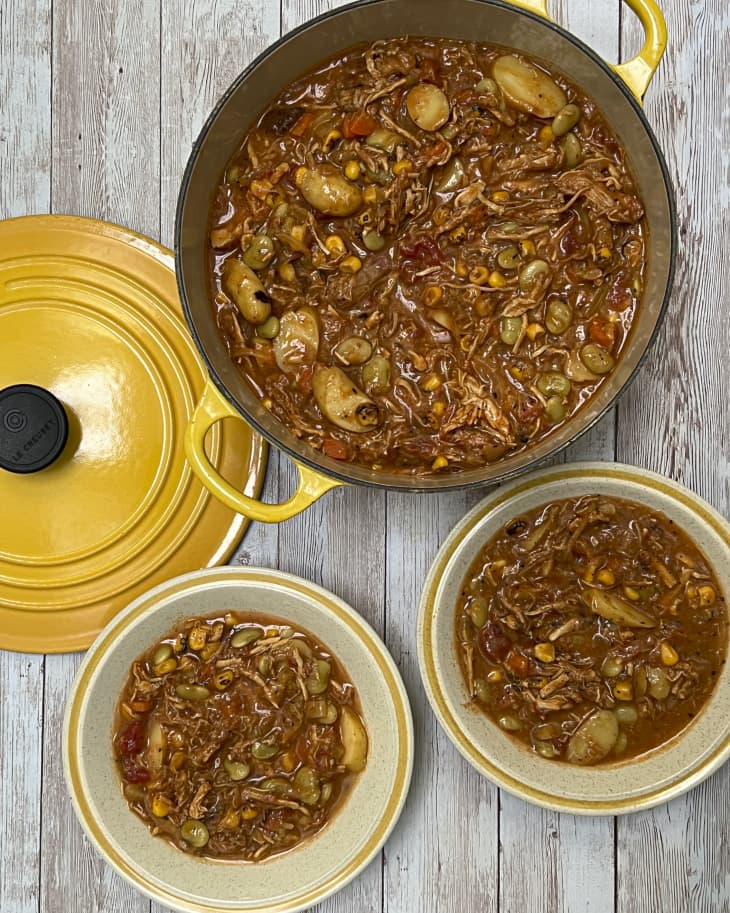 Brunswick stew (a Southern dish that features a tomato base with beans, vegetables, and meat) in a cooking pot with yellow handles, with a yellow lid on the side, and two bowls with stew in them, seen from the top down